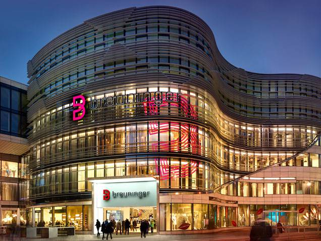 Fassade des Breuninger Flagship‑Hauses in Düsseldorf am Abend, verglaster Eingangsbereich mit rotem Markenlogo und beleuchteten Schaufenstern