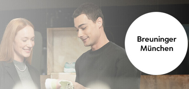 Zwei Personen stehen im Gespräch in einem Modegeschäft; rechts ein runder Hinweis mit der Aufschrift „Breuninger München“, Verkaufsfläche und Kund:innen unscharf im Hintergrund.