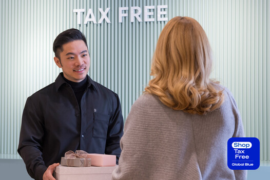 Ein Breuninger-Mitarbeiter übergibt einer Kundin an einem „Tax Free“-Schalter ein Geschenkpaket. Im Hintergrund eine Wand mit geriffelter Struktur.
