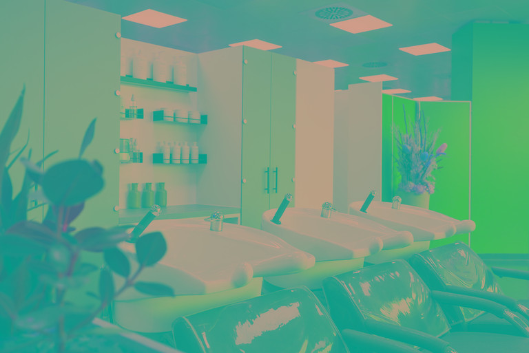 Moderner Friseursalon mit vier weißen Waschbecken und schwarzen Liegesesseln. Dahinter Regale mit Pflegeprodukten und grauen Schranktüren. Links vorne eine grüne Pflanze, rechts hinten ein Blumenarrangement.