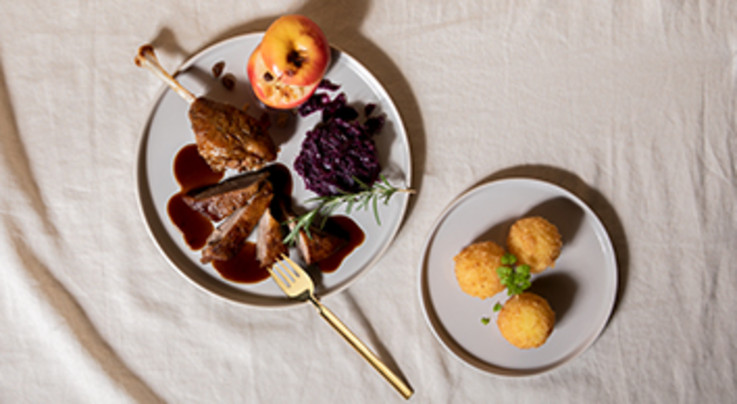 Auf dem Bild ist ein stilvoll angerichtetes Gericht zu sehen, das aus klassischen Komponenten eines Festtagsessens besteht. Im Vordergrund liegt eine Portion Entenbraten, serviert mit dunkler Sauce, begleitet von Rotkohl und einem Bratapfel. Daneben auf einem separaten Teller befinden sich drei goldbraune Kartoffelknödel, garniert mit einem kleinen Zweig frischer Kräuter. Die Farben und Texturen harmonieren perfekt und vermitteln ein Gefühl von Gemütlichkeit und Genuss.