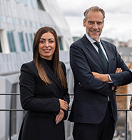 Drei Geschäftspersonen in formeller Kleidung stehen lächelnd auf einer Terrasse mit moderner Architektur im Hintergrund.
